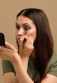 Une personne applique un eyeliner bleu-vert avec un crayon noir sur un fond neutre, reflétant la lumière sur une peau lisse et claire.