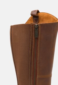 Gros plan de la partie supérieure d'une botte en cuir marron avec fermeture éclair latérale et panneau élastique sur un fond clair.