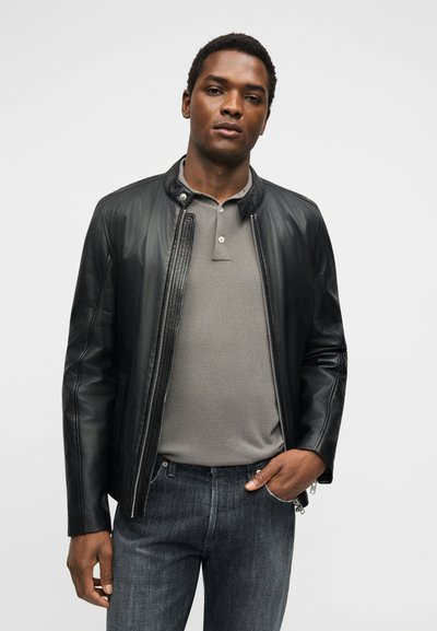 Νέος άντρας με κοντά μαλλιά που φοράει μαύρο δερμάτινο μπουφάν πάνω από γκρι πόλο και σκούρα τζιν, στέκεται με το ένα χέρι στην τσέπη.