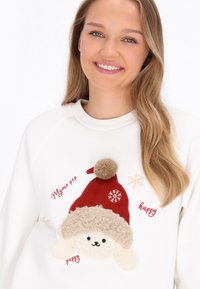 Fehér pulóver piros filc Mikulás sapkával és applikált, krém színű, bolyhos bundájú kiskutyával. Vörös színű szöveges elemek, köztük hópelyhek.