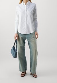 Camicia bianca con bottoni e maniche lunghe, abbinata a jeans chiari a gamba larga e una piccola borsa di denim, completata da sandali neri con tacco.
