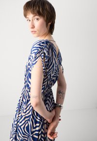 Robe à motifs zébrés bleus et beiges avec des manches courtes, taille froncée et détails à nouer sur les manches. Le modèle a des tatouages visibles sur les bras.
