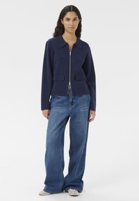 Marinblå zip jacka med krage, två framfickor och långa ärmar, tillsammans med vida blå denimjeans och ljusa skor.