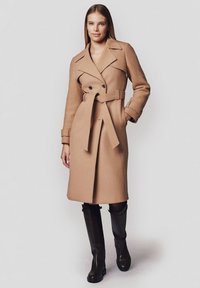 Płaszcz typu camel z podwójnym zapięciem na guziki, paskiem w talii i klapami. Gładki materiał o dopasowanym kroju, zestawiony z botkami do kolan.