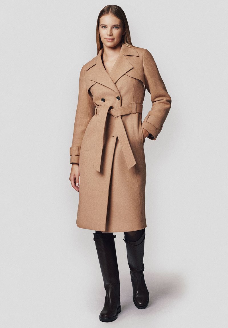 Płaszcz typu camel z podwójnym zapięciem na guziki, paskiem w talii i klapami. Gładki materiał o dopasowanym kroju, zestawiony z botkami do kolan.