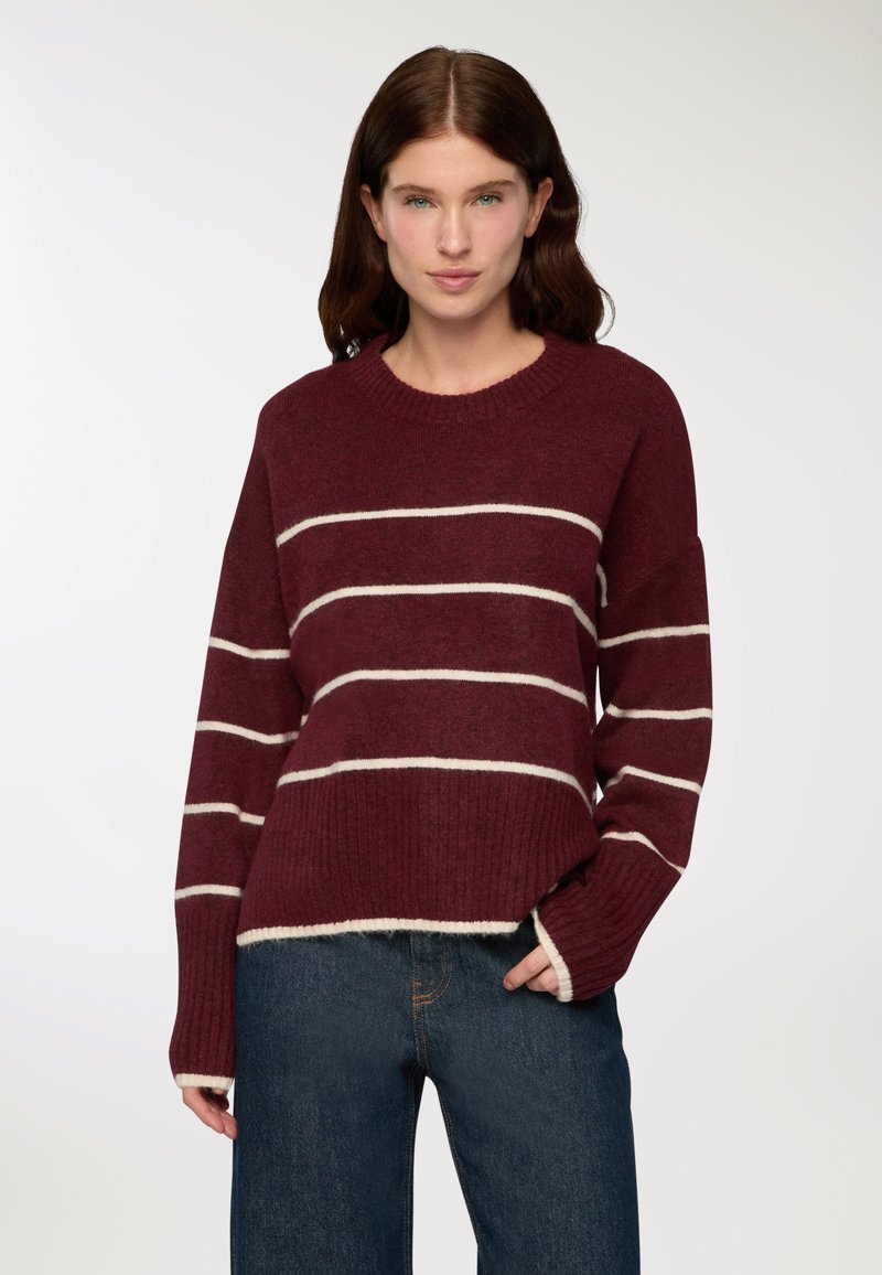 Kobieta z długimi brązowymi włosami nosi bordowy sweter w białe poziome paski i niebieskie dżinsy, stoi z jedną ręką w kieszeni.