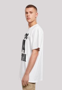 White cotton t-shirt with a loose fit, featuring black printed text on the front. Model wears khaki pants. Smooth texture and short sleeves.