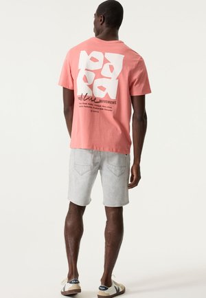 Rosa Baumwoll-T-Shirt mit großem weißem Grafiktext auf der Rückseite. Kombiniert mit hellgrauen Jeansshorts und beigen Sneakers.