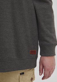 Grauer Pullover mit gerippten Bündchen und Saum, versehen mit einem braunen Lederschild mit "BLEND." Der Arm ist in lässige khakifarbene Hosen gekleidet.