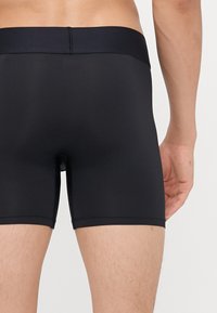 Boxers noirs en tissu lisse et extensible. La ceinture est élastique et se place au-dessus des hanches, avec un logo discret.