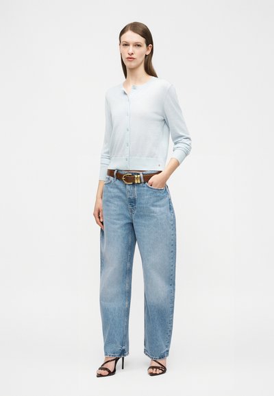 Kvinne står iført lyseblå knappet cardigan, høytlivs løse blå jeans, brunt belte og svarte sandaler med hæl mot hvit bakgrunn.