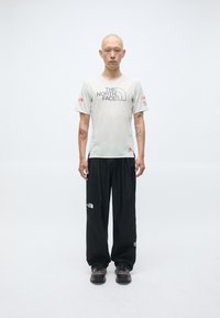 Camisa de manga corta gris claro con logo negro, combinada con pantalones holgados negros y zapatos oscuros con acentos coloridos.