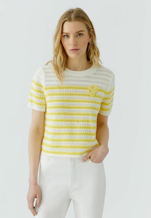 Frau mit blondem Haar, die ein weiß-gelb gestreiftes, gehäkeltes Kurzarmoberteil und weiße Hose trägt und mit einer Hand in der Tasche posiert.