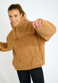 Giacca a pullover in fleece marrone con colletto alto, ampia vestibilità, tasche frontali e tessuto testurizzato. Modella che indossa leggings neri.