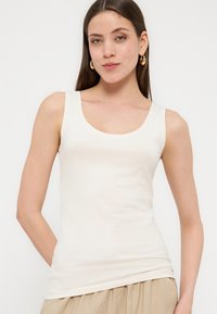 Γυναίκα με μακριά καστανά μαλλιά που φοράει χρυσούς κρίκους στα αυτιά, λευκή αμάνικη μπλούζα και μπεζ παντελόνι, στέκεται με τα χέρια στις τσέπες.