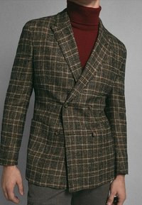 Blazer doppiopetto in tessuto plaid marrone con una sottile texture. Presenta rever larghi e due tasche frontali. Indossato sopra un dolcevita bordeaux.
