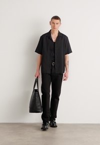 Camiseta negra de manga corta, pantalones ajustados negros, zapatos de cuero negros y un bolso de mano de cuero negro. Collares de cadena de plata y tatuaje visible.
