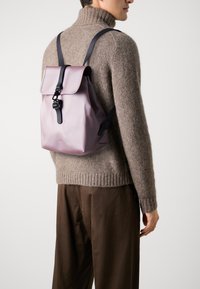 Persona che indossa un maglione a maglia beige e pantaloni marroni, mentre porta sulla schiena uno zaino malva con spalline e fibbia nere.