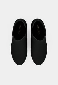 Par de zapatos negros sin cordones con lados elásticos, vistos desde arriba sobre un fondo blanco.