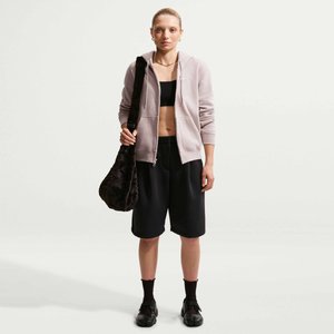 Jeune femme portant un sweat à capuche zippé rose clair, un soutien-gorge de sport noir, un short long noir, des chaussettes noires, des chaussures, et portant un grand sac noir.