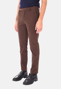 Uomo che indossa pantaloni in corduroy marroni con bottone e tasche laterali, abbinati a scarpe di pelle nere eleganti e una camicia blu navy.