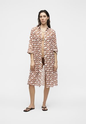 Žena nosiaca vzorovanú hnedo-bielu dlhú košeľu na gombíky cez tmavé spodné prádlo a čierne šľapky, stojaca pred jednofarebným pozadím.