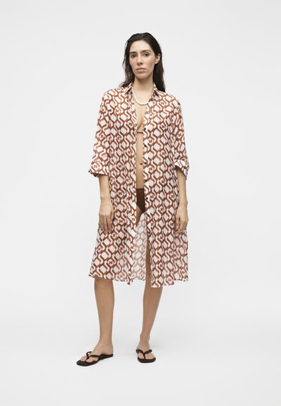 Femme portant une chemise longue boutonnée marron et blanche à motifs par-dessus des sous-vêtements foncés et des tongs noires, debout devant un fond uni.