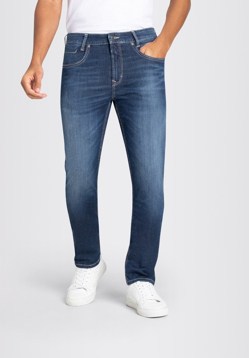 Jeans en denim bleu foncé avec une coupe slim, design à cinq poches et couture visible. Associés à des baskets blanches, portés par une personne.