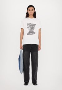Hvid T-shirt med sort grafisk design, der viser en snegl og teksten "MEOW GARY," parret med løse mørke jeans og holder en blå denimjakke.
