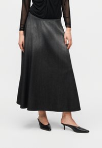 Μακριά γκρι τζιν φούστα με λεία υφή και ροή στη σιλουέτα, σε συνδυασμό με μαύρα μύτα παπούτσια. Η φούστα έχει απλή γραμμή στον ποδόγυρο.