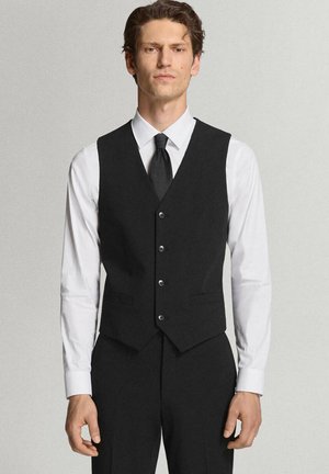 Mann trägt eine schwarze Anzugweste und Hose mit weißem Hemd und schwarzer Krawatte, steht vor einem schlichten hellen Hintergrund.