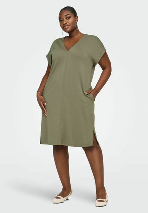 Donna che indossa un vestito verde oliva lungo fino al ginocchio con maniche corte e scollo a V, in piedi con una mano in tasca, ballerine beige e orecchini.