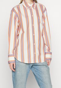 Skjorta med knappar i orange och blå vertikala ränder på en krämfärgad bakgrund. Har en bröstficka och långa ärmar med manschettdetaljer.