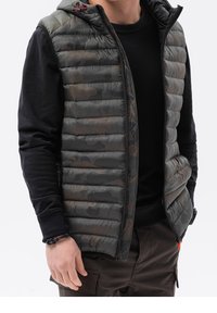 Gepolsterte, kapuzenlose Weste in dunklem Camouflage mit einem schwarzen Langarmunterhemd. Merkmale sind ein Reißverschluss und rote Akzente an der Kapuze.