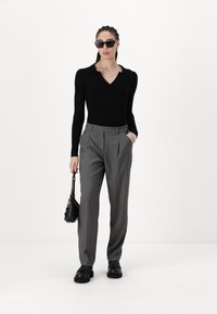 Haut à manches longues noir à côtes avec un col en V, associé à un pantalon gris avec des plis. Les accessoires comprennent des lunettes de soleil et un petit sac à main noir.