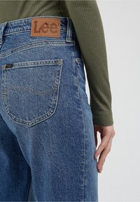Modré džínové kalhoty s uvolněným střihem a tmavým švem, s hnědou koženou nášivkou s nápisem "Lee" na zadním pásu.