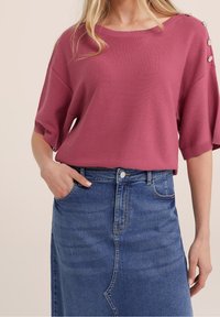 Fuchsiastrumptröja med ribbat mönster och korta ärmar, med knappdetaljer på axlarna, som är ihop med en högmidjad blå denimkjol med en söm fram. 