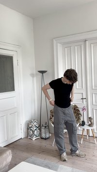 Ikdara melna kreklu, pelēkas kabatbikses ar sānu kabatām un beigu sporta apavus. Iekštelpas ar baltām sienām un koka grīdu.