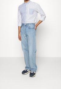 Man som bär en vertikalt randig blå och vit skjorta med knäppning, ljusblå lössittande jeans och blå och vita sneakers, står med handen på höften.