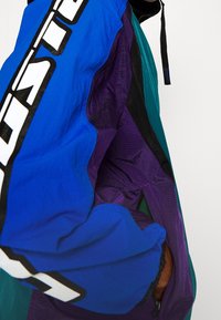 Colorful windbreaker featuring blue, purple, and green panels. Textured, lightweight fabric with a bold logo on the sleeve and adjustable cuffs.