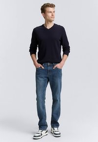 Marinblå v-ringad tröja, ljusblå jeans och vita sneakers med svarta detaljer. Modellen står med händerna i fickorna, neutral bakgrund.