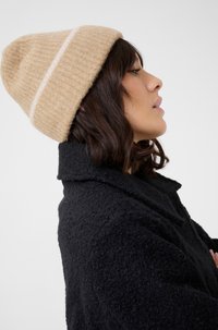 Berretto a coste di colore crema con una striscia bianca a contrasto, indossato con un cappotto nero testurizzato. Vista di profilo, che evidenzia il materiale e il design.
