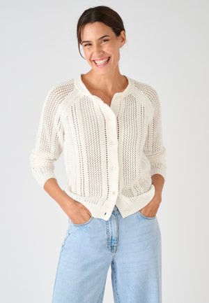 Vrouw glimlachend, gekleed in een witte gebreide cardigan met knopen en lichtblauwe spijkerbroek, met handen in de zakken tegen een effen achtergrond.