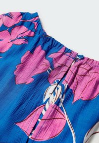 Vêtement bleu à large imprimé floral rose avec un col froncé comportant un petit bouton bleu et une fermeture à œillet.