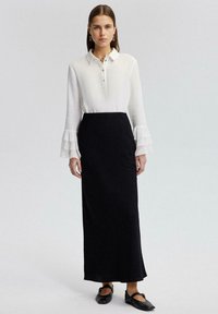 Camisa blanca de cuello con puños acampanados, combinada con una falda larga negra ajustada. El modelo lleva zapatos planos negros, que presentan un diseño elegante.