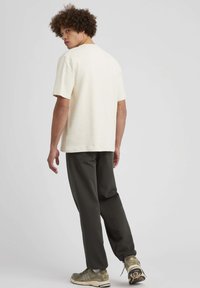 Chemise beige clair à manches courtes avec un tissu texturé, associée à un pantalon vert foncé et des baskets gris-brun. Le modèle montre la vue arrière.