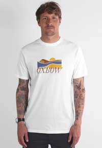 Homme avec des tatouages sur les deux bras portant un t-shirt blanc avec un paysage abstrait coloré et le texte "OXBOW", se tenant devant un fond uni.