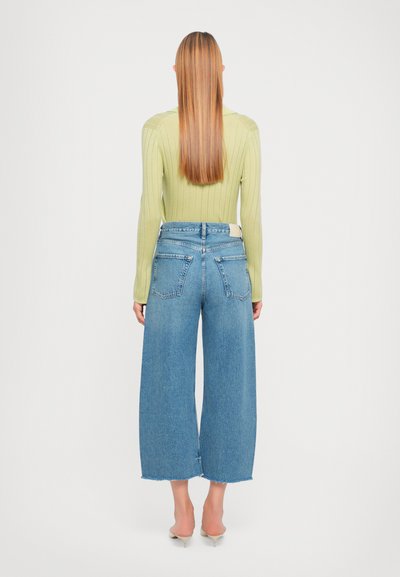 Pull à côtes vert clair associé à un jean en denim bleu à jambe large avec des ourlets bruts et une conception à cinq poches, vu de dos.