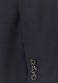 Manche de costume bleu marine foncé avec trois boutons sombres disposés verticalement près du poignet sur un tissu texturé.