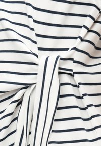 Λευκό ύφασμα με ομοιόμορφες οριζόντιες μαύρες ρίγες, δεμένο σε έναν χαλαρό κόμπο, που δείχνει την υφή και την ελαστικότητα του υλικού.